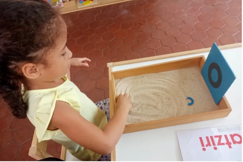 A child uses a sand tray to practice tracing letters, a tactile activity that prepares the hand for writing.